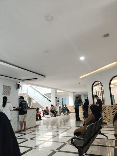 The image depicts a lobby area inside a building located at coordinates -6.32 latitude and 107.293 longitude in Telukjambe, Indonesia. The lobby has a white tiled floor, white walls, and a white ceiling with recessed lighting fixtures. There are several people waiting in the lobby. Three people are standing in line at a marble-topped counter. Two people are seated on a bench, while another two people are seated on chairs. There is also one person in a wheelchair. Two people are standing nearby, and one is seated. There is a moving staircase on the left side of the lobby. The entrance is at the back of the lobby. There is a large window with a decorative metal grill behind the counter.