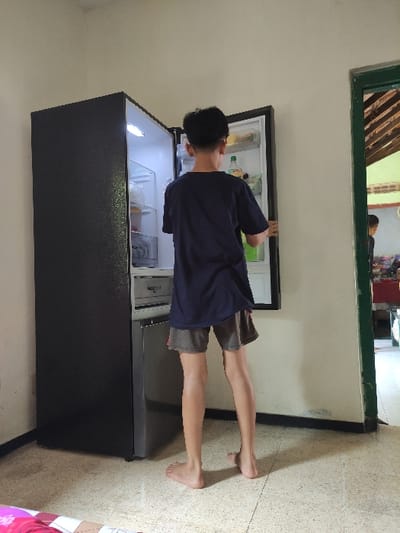 A young man, wearing a dark blue t-shirt and khaki shorts, is standing in front of a refrigerator in a kitchen. The man is looking at something inside the refrigerator, which is open, and is using his right hand to hold the refrigerator door. The kitchen has white walls and a light brown tiled floor. The refrigerator is black and has a silver freezer section. The scene is located in Kecamatan Lawang, Indonesia, at coordinates: -7.828 latitude, 112.701 longitude. There is a door leading to another room in the background of the image. The door is green and has glass panes, allowing one to see a portion of another room with a white tiled floor and walls. A person is standing near a table in the other room. The kitchen is minimally furnished.