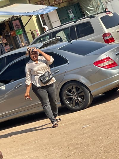 The image captures a daytime street scene in Samaru, Nigeria. A woman stands in the foreground, slightly left of center, shielding her eyes with her raised right hand. She is dressed in a light-colored, long-sleeved collared shirt, dark jeans, and black open-toed sandals. A black quilted shoulder bag with a chain strap is worn across her body. Her left hand holds a small object with a red accent. Behind the woman, a silver Mercedes-Benz C300 sedan is parked, featuring black multi-spoke alloy wheels. To the rear of the Mercedes, a light-colored SUV, identifiable as a Toyota Highlander by its rear design, is also parked. In the background, storefronts are visible; one features a blue and white striped awning covering shelves stocked with bottled and packaged goods, and a poster on its wall. Adjacent to it, another structure has a dark green roll-up door with a "POS" sign. A person wearing a white head covering is partially visible behind the SUV. The ground beneath the subjects and vehicles appears to be unpaved dirt or gravel. The scene is brightly illuminated by direct sunlight, casting distinct shadows.