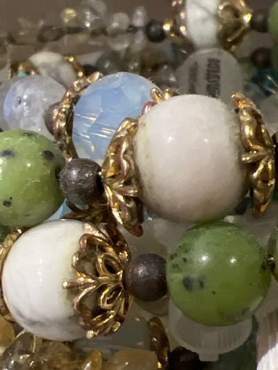 The image displays a close-up, macro shot of a beaded bracelet. The bracelet is composed of various beads, including round white beads, green speckled beads, and a faceted light blue bead. There are also dark, metallic-looking beads and ornate gold-colored bead caps. The background is blurred, showing more beads and possibly other jewelry. There are no people or any recognizable elements of the environment visible, making it impossible to determine a location in Sinyavino, Russia, or any other geographical context.