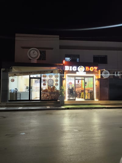 This is a nighttime, eye-level, medium shot of two storefronts on a street in an urban area, likely in Tunisia. The storefront on the left is for "PATISSERIE LA PERGOLA" and displays images of pastries and bread. The storefront on the right is for "BIG BOY TACOS" and features a brightly lit entrance and seating visible inside. Both establishments are illuminated, and the street in front is wet, reflecting the lights. The background is dark, suggesting it is nighttime.