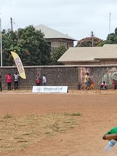 An outdoor scene in Waa Ngombeni ward, Kenya, depicts a reddish-brown dirt ground with sparse, dry grass patches in the foreground. A blurry figure in a green top holding a water bottle is partially visible in the bottom right, facing away.
Along the midground, a dark brick wall extends horizontally, displaying faded paint and graffiti on its right side. Against this wall, from left to right: a tall, yellow "Betika" banner stands vertically. To its right, two adult males are standing; one wears a red shirt with white sleeves, the other a black and white patterned shirt. A white horizontal banner featuring "BingwaFest" and a logo rests on the ground in front of the wall. To the left of this banner, a male in a green shirt and red cap holds a red item while another person in a white shirt is partially visible behind him. Further right, a male in a yellow shirt rides a yellow bicycle with an indistinct seat. Adjacent to him, a male in a red and white shirt is seated on the ground. Several children are gathered on the far right, some standing, some seated.
Behind the wall, green trees with dense foliage are visible. Several buildings with light-colored corrugated metal roofs and exposed stone or concrete block walls are visible, particularly on the left and center. Utility poles and overhead lines are present against an overcast, light grey sky. The scene is illuminated by diffused daylight.