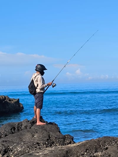 The image depicts a person fishing off a rocky shore in Canggu, Indonesia. The individual, who is wearing a black full-face helmet and a face covering, stands barefoot on a dark, porous rock formation. They are dressed in a light-colored hooded sweatshirt and dark shorts, with a black backpack slung over their shoulder. The person holds a fishing rod with a visible reel, casting a line out into the blue ocean, which stretches to the horizon under a sky with scattered white clouds. Other dark rocks are visible to the left of the person. The location can be presumed to be a coastal area in Canggu, given the rocky shoreline and ocean view.