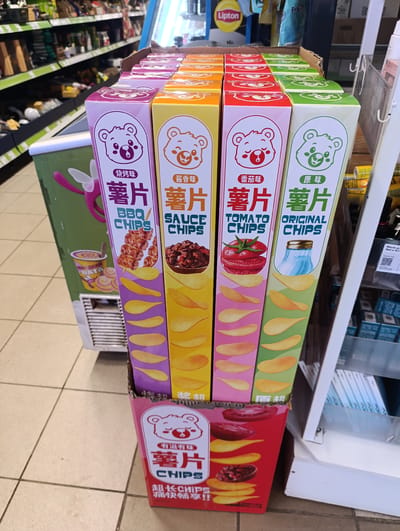 The image shows a display of Chinese potato chips in a store. There are four different flavors available: BBQ, Sauce, Tomato, and Original. Each flavor comes in a tall, rectangular box with a cartoon bear character and images of the chips and corresponding flavor ingredients. The display is in the aisle of a grocery store, likely in Fryanovo, Russia, given the OCR content that appears to be in Russian. Other grocery items are visible on shelves in the background. The display is located adjacent to a beverage cooler and a display stand.