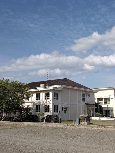 The image depicts a low-rise, light-colored building with a dark, sloped roof, situated in Mesjid Reuntoh, Indonesia. The building has a modern architectural style with numerous windows. In front of the building, several parked cars and motorcycles are visible. A large tree with dense green foliage is on the left side of the frame, and a smaller tree is in front of the building. The ground in the foreground is covered with small, light-colored gravel. The sky above is blue with scattered white clouds. A small, open-sided structure with a corrugated metal roof and a cage-like enclosure stands near the building. A water tank is also visible on the roof of the main building. Another building, lighter in color, is partially visible to the right.