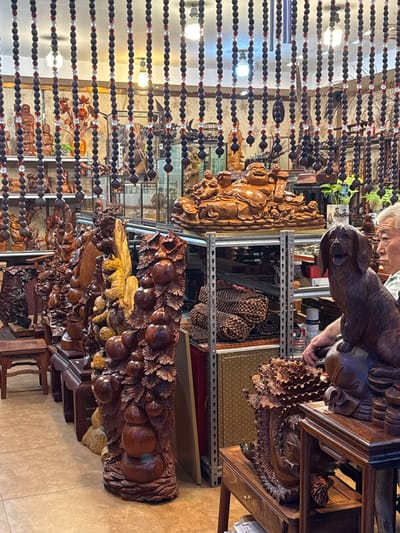 The image depicts a densely packed display of wooden carvings inside a shop, likely in Linghe, China. The shop appears to be a retail environment specializing in decorative art objects made from wood. The items range from large, intricate sculptures to smaller decorative pieces. In the foreground, there is a large wooden carving of a dog, with a man's arm resting on its head. Behind the dog carving, shelves are filled with various wooden items, including what appear to be stacks of round, carved objects and other figures. Further back, a prominent carving of a laughing Buddha on a tiered base is visible, surrounded by other smaller wooden figures and decorative elements. The shop's interior is further enhanced by strings of wooden beads with red and clear accents hanging from the ceiling, creating a curtain-like effect. The lighting suggests an indoor setting with overhead illumination. The overall impression is of a marketplace or a store dedicated to the sale of traditional Chinese wooden crafts.