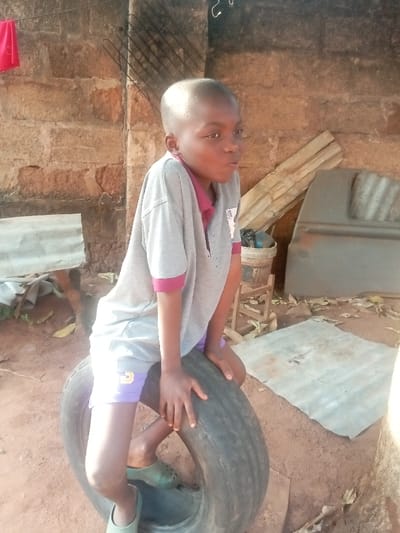 In Ogidi, Nigeria, a young male child is seated on an upright black tire outdoors. He wears a grey short-sleeved polo shirt with maroon collar and sleeve cuffs, light purple shorts with a yellow 'S' or '5' emblem on the left thigh, and light green slip-on shoes. The child, with a closely shaven head, faces slightly right, his mouth partially open, and both hands rest on the top edge of the tire. The background features a rough, unpainted wall constructed from reddish-brown brick or block. The ground consists of exposed reddish-brown earth. Visible background objects include a section of corrugated metal to the left, a black metal grate and a red fabric item affixed to the wall higher up. To the right, several wooden planks lean against the wall, alongside an overturned bucket, a basic wooden stool, and a large, dark, curved metal panel, possibly a vehicle component. Another flat piece of corrugated metal and scattered organic debris are present on the ground. The scene is illuminated by natural daylight.