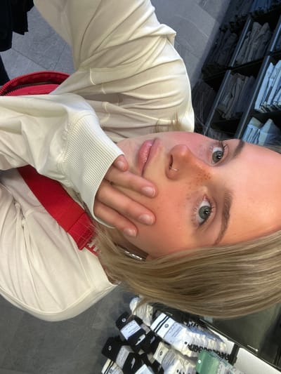 A close-up selfie of a fair-skinned, blonde-haired woman with freckles and green eyes is taken indoors, angled from above and slightly off-center. She wears a light-colored, long-sleeved sweater with a ribbed cuff and a red strap or bag is visible on her shoulder. Her right hand partially covers her face. The background shows shelves of clothing or merchandise, and stacks of socks packaged with the Lululemon logo. The photo appears to be taken inside a Lululemon store, possibly located in the city of Whistler, Canada. The floor is a dark gray tile.