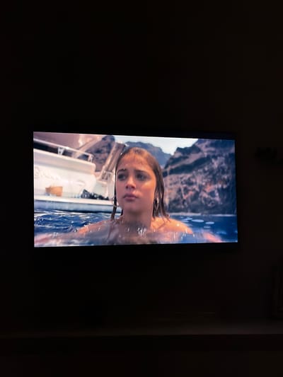 This is a vertical, medium shot of a young woman in the water, likely a pool or a lake, seen from the chest up. She has long, light brown hair that is wet and clinging to her face and shoulders. Her eyes are open and looking directly at the camera. She appears to be emerging from or interacting with the water, as evidenced by the droplets and reflections on her skin and hair. The water is clear and blue, with ripples indicating movement.
In the background, there's a hint of a white structure, possibly a boat or a poolside amenity. Beyond that, a rocky, mountainous landscape is visible, bathed in what appears to be natural light, suggesting it's daytime. The overall mood is serene and perhaps a little mysterious, given the close-up on the woman's face and the surrounding environment. There is no visible text in the image. The location context provided is Sofiivska Borshchahivka, Ukraine.