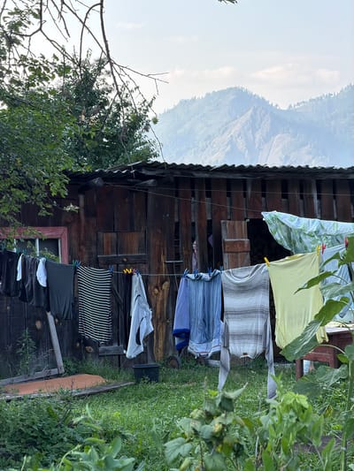 The image depicts an outdoor scene at what appears to be a rural dwelling in Anos, Russia. A wooden structure, possibly a shed or outbuilding, with a corrugated metal roof, is centrally located. Clothes are hanging on a clothesline in front of the structure. The clothes include various garments such as t-shirts, trousers, and what looks like a striped dress or skirt. Some clothes are secured with clothespins. In the foreground, lush green grass and foliage are visible, with some taller plants, possibly sunflowers, to the right. In the background, a hazy view of mountains is seen under a sky with scattered clouds. The overall atmosphere suggests a mundane, everyday scene in a remote, natural setting. There are no people visible in the image.