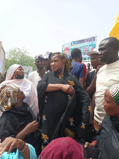 This outdoor photograph captures a daytime gathering of people, predominantly adults, under a bright, clear sky in Giginyu, Nigeria. The scene suggests a political event, characterized by a large banner visible in the background. The banner prominently displays "COMMANDER" in bold lettering, alongside "Vote First APC," and features headshots of several men, including those identified as "Haruna Jowa" and "Murtala Gwa," presumably political figures or candidates.
The foreground is filled with a diverse group of individuals. Several women are visible, many wearing traditional attire such as hijabs and patterned headscarves. One woman in the center wears a black garment adorned with distinctive yellow and orange embroidered patterns and a black face mask. To her left, another woman in a light-colored hijab and a black face mask looks upwards. Other women wear dark clothing and colorful head wraps, some accessorized with bracelets and earrings.
Among the men, some wear traditional caps and tunics, while others are dressed in more casual attire like t-shirts and baseball caps. They are standing in close proximity, engaged in observation or conversation. A partial yellow flag or fabric is visible on the far right of the frame, and a wooden frame of another sign is on the far left. The overall atmosphere indicates a public assembly or rally.