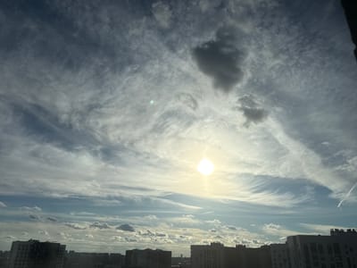 The image captures a broad view of the sky over Lyubertsy, Russia, featuring a mix of cloud formations and the sun. In the lower portion of the frame, the silhouettes of several multi-story buildings are visible, indicating an urban setting. The sun is a bright, diffused disc in the upper center of the sky, surrounded by a halo of light. Wispy, cirrus-like clouds stretch across the sky in various directions, while some scattered cumulus clouds are also present, particularly lower in the scene. The overall lighting suggests it is daytime, possibly late afternoon given the angle and intensity of the sun. No individuals or specific actions are discernible in the image.