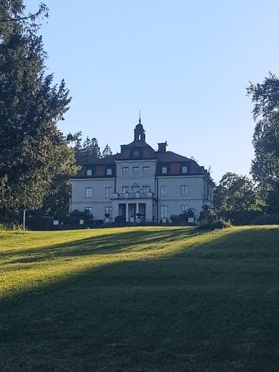 The image shows a large, pale grey manor house with a red-tiled roof and a prominent dark-capped cupola on top, situated in a rural setting. The house has multiple windows and a balcony supported by columns. It is surrounded by lush green trees, some of which are tall evergreens. The foreground is a sloping, sun-drenched lawn with distinct shadows cast across it, suggesting it is late afternoon or early morning. The sky is clear and blue. The manor house is located in Skriketorp, Sweden, a historical estate with a parkland setting.