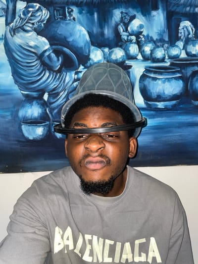 The image presents an indoor, chest-up portrait of an adult male looking directly forward. The male has dark skin, short dark hair, a mustache, and a goatee. He exhibits a facial expression with pursed lips and slightly narrowed eyes. On his head, he wears two stacked transparent plastic objects: a translucent, textured plastic bowl on top, and a clear plastic ring positioned around his forehead, partially obscuring his upper eyes. He is wearing a grey long-sleeved t-shirt with "BALENCIAGA" printed across the chest in distressed light-colored lettering. The background is dominated by a large, monochromatic painting rendered in shades of blue and indigo. This painting depicts multiple figures, seemingly engaged in tasks involving large pottery vessels, with a prominent figure on the left carrying a substantial pot. The environment appears to be an interior space with a light-colored wall. The specific geographic location within the city of Ube, Nigeria, cannot be determined from the visual information present in the image.