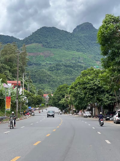 The image depicts a street scene in Hà Giang 2 Ward, Vietnam, characterized by a paved road lined with trees and buildings. The road runs through a valley with lush, green, heavily forested mountains dominating the background. The sky is overcast with heavy clouds.
On the left side of the road, there are a few buildings with red roofs, interspersed with banana trees and a large banner displaying Vietnamese text and images. Electrical poles and wires are visible along the roadside. Several parked motorcycles and scooters are present. A person on a white scooter, wearing a green jacket and helmet, is riding towards the viewer. Further down the road on the left, there are some stalls or makeshift shops covered with umbrellas.
The road itself is a two-lane asphalt surface with yellow and white lane markings. Several vehicles are present on the road. A dark-colored SUV is driving away from the viewer. On the right side of the road, there are more trees and buildings. A person on a black scooter, wearing a dark jacket and helmet, is riding away from the viewer. Parked cars, including a white sedan and a black SUV, are visible along the right curb.
The overall impression is a moderately busy street in a semi-rural or suburban area, nestled within a mountainous landscape under cloudy weather.