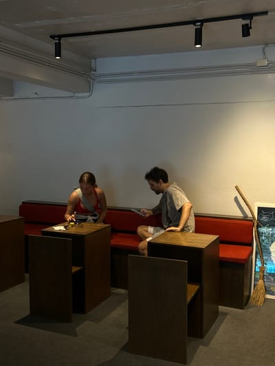 The media file shows two individuals in a dimly lit indoor setting that appears to be a cafe or a communal workspace. The scene is oriented sideways, with the ceiling running vertically on the left and the floor horizontally at the bottom.
The subjects are a man and a woman. The man, wearing a grey t-shirt and shorts, is bent over a wooden table, looking down at something in his hands, possibly a phone or a device. He has dark hair and a mustache. The woman, wearing a red top and grey straps, is also focused on a table, her hands engaged with an object on the surface. She has dark hair tied back.
Several wooden tables of varying sizes and configurations are visible, some appearing as modular units. A red bench is situated behind the man. In the background, a broom with a straw head and a wooden handle leans against the wall above the red bench. The walls are painted in light and darker tones, with electrical conduits and junction boxes visible on the lighter section. Two black spotlights are mounted on a vertical pole on the left side of the frame, with one emitting light.
The overall atmosphere suggests a casual, perhaps late afternoon or evening setting, given the indoor lighting. There is no visible text in the image. The location context of Chiang Mai City Municipality, Thailand, is provided, but no specific visual cues within the image definitively confirm this location.