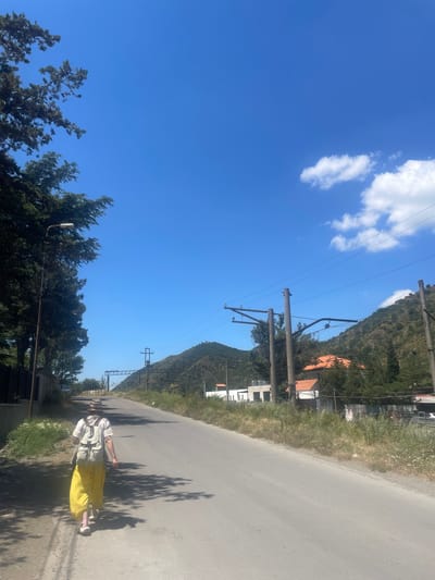 A person wearing a straw hat, white shirt, and yellow pants walks away from the camera down a paved road in Mtskheta, Georgia. The person carries a light-colored backpack. The road is bordered by grassy verges and trees on the left, and more trees and buildings on the right. Hills covered in vegetation rise in the background, with power lines and poles visible against a clear blue sky with scattered white clouds. The scene appears to be during daylight.