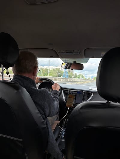 The image shows a car interior with a driver. The driver is an older man wearing a gray sweater and glasses, holding the steering wheel. Two smartphones are mounted on the dashboard. One displays a map and the other is likely a rideshare app screen. The scene is set in Kuznechiki, Russia, indicated by the view through the windshield which shows a city street and residential buildings. A railway bridge can be seen on the left side of the frame.