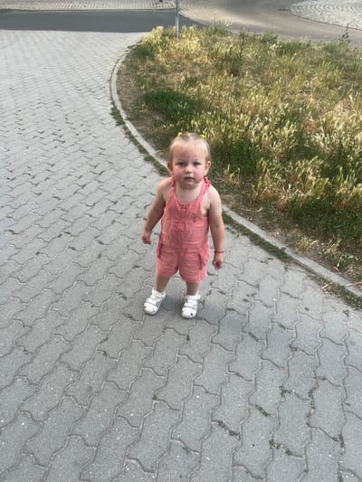 A young child stands on a grey interlocking paver pathway in Pilsen, Czechia. The child, wearing a pink romper and white sandals, has blonde hair styled with yellow hair ties. To the right of the pathway, a patch of dry, tall grass is visible. The pathway curves slightly to the left, with a dark grey paved area beyond it. The image is taken from a slightly high angle, looking down at the child.