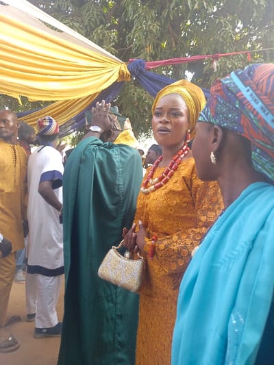 The image shows a group of people outdoors, possibly at a wedding or celebration. A woman in a yellow lace dress, head wrap, and red beaded necklace holds a silver purse and appears to be speaking. Another woman with a headscarf is beside her, partially obscuring the view. Behind them, people are dressed in traditional attire, including green robes and white outfits with blue trim. Yellow and blue fabrics hang overhead.