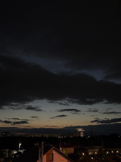 The image depicts a twilight scene over the city of Kondratovo, Russia. The sky is dominated by heavy, dark clouds, suggesting impending rain or a storm. Below the clouds, a gradient of light transitions from deep blue and gray in the upper sky to a faint orange and yellow glow near the horizon. A line of buildings, illuminated by lights, stretches across the middle ground, appearing as a silhouette against the dimly lit sky. Among these buildings, one prominent structure stands out with a series of bright lights outlining its upper floors, possibly indicating an office building or residential complex. In the foreground, the rooftops of several houses are visible, with some lights on within the structures. The overall atmosphere is one of a darkening evening in a suburban or semi-urban environment. No people or specific actions are discernible in the image.