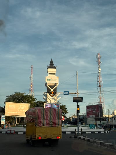 The image shows a street scene in Lambaro, Indonesia, with a yellow truck partially obscuring the view. A tall BSI (Bank Syariah Indonesia) building is visible, along with telecommunication towers in the background. The sky is cloudy. Street signs and billboards for brands like "Gudang Garam Merah" are present. A traffic light shows a yellow light. There is a blue sign that says "DILARANG MENEROBOS"
---