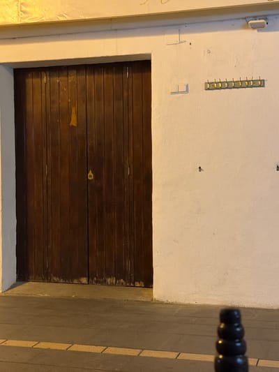 The image shows a closed wooden double door in a white stucco wall. The door has vertical planks and a brass handle with a keyhole. To the right of the door is a light switch and a row of coat hooks with yellow tags. There is also an outdoor light fixture on the wall above the light switch. The ground in front of the door is paved with concrete and there is a yellow line on the pavement. In the foreground, a black bollard is visible. The scene appears to be an exterior of a building in Ibiza, Spain, possibly a residential or commercial property.