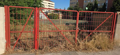 A red metal gate, consisting of two sections, is in the foreground, partially obscured by dry, tall grass and weeds. The left section of the gate is attached to a beige concrete post on the far left. The gate's frame and crossbars are made of red painted metal tubing and wire mesh. A handle mechanism is visible on the left gate section. The right section of the gate is closed and secured with a chain that is attached to a hook on the gate post. Behind the gate, a chain-link fence encloses an area with a dirt surface and some scattered greenery. In the background, a multi-story apartment building with a light-colored facade and a brick building are visible. Trees and bushes are present in the middle ground and background. The sky is clear and blue. The scene appears to be an outdoor urban environment. The location is identified as the city of Huesca, Spain.