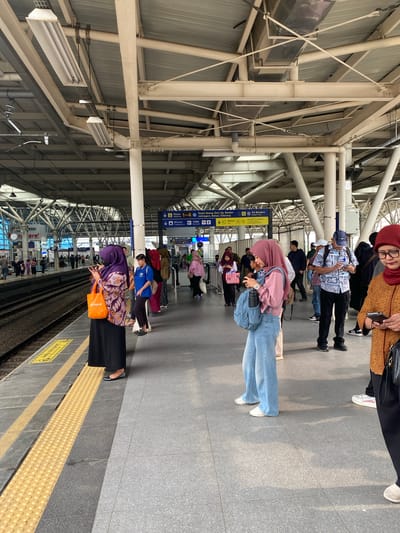 The image depicts a busy train platform at Manggarai Station in South Jakarta, Indonesia. Several individuals, predominantly women wearing hijabs, are standing on the platform, many of them looking at their mobile phones. One woman in the foreground, wearing a purple hijab and a patterned top, holds an orange bag. Beside her, another woman in a pink hijab and light blue jeans is also engrossed in her phone, with a backpack at her feet. A man in a patterned shirt and a blue cap stands further down the platform, also looking at his phone. The platform itself is paved with grey tiles, with a textured yellow safety strip running along the edge closest to the train tracks. Overhead, a complex network of steel beams and light fixtures supports the station's roof. Signs indicating platform numbers and destinations are visible in the background. The scene appears to be a typical moment of transit, with people waiting for their trains.