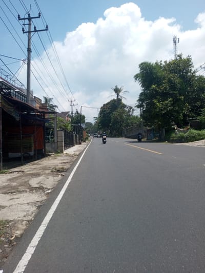 The image depicts an asphalt road in Nawng Hkar, Myanmar. A white line marks the road's edge on the left side. A motorcycle is visible in the center of the road, moving away from the camera. Another motorcycle appears further in the distance, and a white car is visible beyond that. Overhead power lines stretch across the scene. Buildings and trees flank both sides of the road. The sky is blue with scattered white clouds. A cell tower is visible in the background.