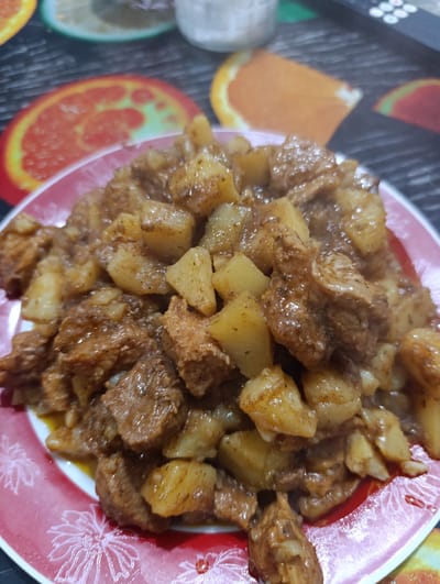 A close-up, overhead shot showcases a red and white plate filled with a hearty dish of cubed potatoes and meat in a rich, brown sauce. The food is piled high, obscuring the plate's design, which features an outline of stylized flowers on the red rim. The background is blurred, but appears to be a tabletop with a printed pattern of fruit slices, possibly oranges and watermelons, and a black surface. The lighting is warm and slightly diffused, suggesting an indoor setting. No people are visible. The location is specified as Kyustendil, Bulgaria.
