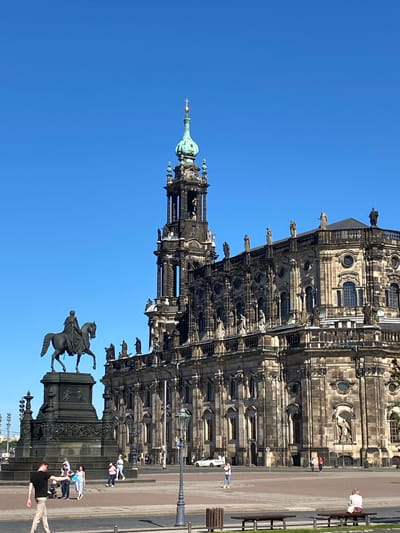 The image depicts the Hofkirche (Dresden Cathedral) and the equestrian statue of King John of Saxony, located in Dresden, Germany. The perspective is from ground level looking upwards and slightly rotated. The Hofkirche is a large baroque-style cathedral. The statue is prominently visible in the foreground. Several pedestrians are walking along the plaza. The sky is clear and blue. Benches are visible near the cathedral. A lamppost is seen on the right. A person is gesturing upwards towards the statue.