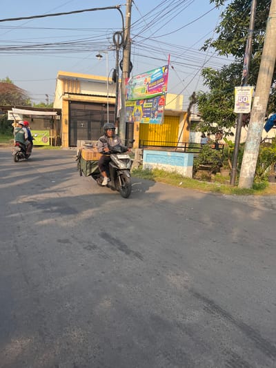 The image depicts a street scene in Karangtanjung, Indonesia, under a clear sky. In the foreground, a man wearing a grey jacket and helmet rides a dark-colored scooter with a large, olive-green cargo bag and a cardboard box strapped to the back. The scooter has a license plate that reads "L 6531 MA". Further down the road, another scooter is visible, carrying two people, one wearing a red cap.
The background reveals buildings with yellow and orange facades, some with large black metal gates. A bright yellow roll-up door is also visible. Utility poles with a tangle of electrical wires dominate the upper portion of the image. Several banners and posters are attached to these poles, including one with text that appears to advertise for a school and another with the number "50" in a red circle. A small, overgrown area with plants and a concrete structure with blue decorative elements is present on the right side of the road. The road surface is asphalt, showing signs of wear and tire marks. The overall impression is of a typical day on a moderately busy street in a residential or commercial area.