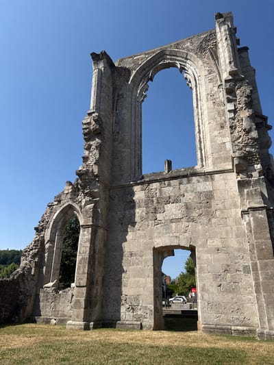 This is a low-angle, full-shot, outdoor photograph of the exterior ruins of the Walkenried Abbey in Walkenried, Germany. The ruins are made of light-colored stone and are crumbling, with large openings in the walls and roof. The sky above is clear and blue. In the foreground, there is a patch of dry grass. Through one of the archways in the ruins, a white car and some buildings can be seen in the background. The image captures the historical and weathered nature of the abbey ruins.