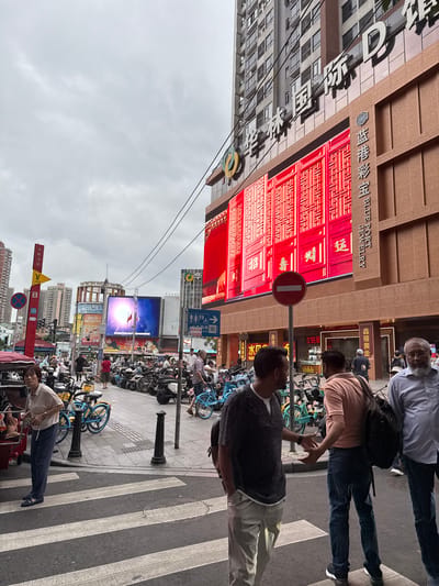 A street scene unfolds in Liwan District, China, under an overcast sky. The foreground features a pedestrian crossing with white stripes on asphalt. To the left, a woman stands near a red vehicle, wearing a light-colored short-sleeved shirt and dark pants. In the mid-ground, three men are visible. The leftmost man, wearing a dark, textured v-neck sweater over a white t-shirt and light-colored pants, gestures with his left hand. Next to him, a man in a peach-colored t-shirt and dark jeans faces away, carrying a black backpack. To the right, a man with a beard and glasses, dressed in a light blue button-down shirt and dark pants, stands looking towards the left. A row of parked motorcycles and bicycles stretches across the mid-ground. Behind the pedestrians and vehicles, large digital billboards display vibrant red graphics and text, including Chinese characters and the words "BLUE PORT JEWELRY." A tall, modern building with many windows rises behind the billboards. A red traffic sign with a white horizontal bar is also present. The overall impression is a busy urban environment with commercial activity.
