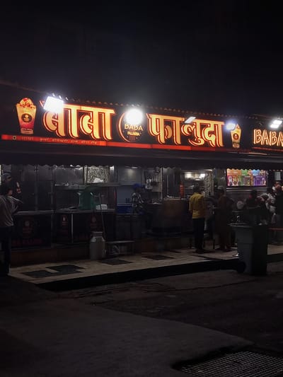 The image presents a nighttime street scene in Mumbai, India, dominated by a brightly lit shop named "Baba Falooda." A large, vertical neon sign, glowing in yellow and red, prominently displays "बाबा फालुदा" in Hindi and "BABA FALOODA" in English. The sign also features a graphic of a dessert cup with "ROYAL MALAI" written on it. Further down the sign, in Hindi, are listed several branch locations: "शाखा :- कुर्ला, जोगेश्वरी, ठाणे, मीरा रोड." (Branches: Kurla, Jogeshwari, Thane, Mira Road.), confirming its presence in the Mumbai and Thane regions.

Below the illuminated sign, the shop appears to be a lively establishment. Several individuals, likely customers and staff, are visible, gathered around counters. One person in a yellow shirt is noticeable near the serving area, suggesting active business operations. The shop's setup includes various display units and what seem to be commercial refrigerators or counters. To the right side of the shop, two gas cylinders are stacked against a wall. The ground is paved, and a dark metal grate is visible on the far right. The overall atmosphere indicates a popular urban eatery open for business after dark, with artificial lighting providing the primary illumination against the night sky.