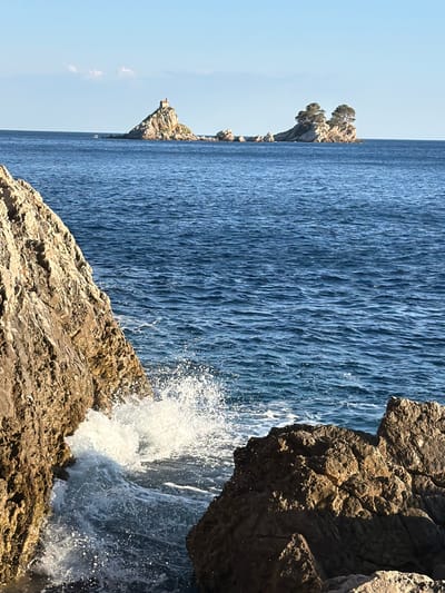 The image depicts a coastal scene with the sea in the foreground and middle ground, and several small islands in the distance. In the immediate foreground, rough, textured rocks are visible on the left and right, with waves crashing between them, creating white spray. The sea is a deep blue with moderate ripples, reflecting the clear sky above. In the distance, a cluster of rocky islands emerges from the water. One island in the center-left has a small building with a red roof on its peak. To the right of this, another larger island is topped with several evergreen trees. A small white boat can be seen on the water to the far left, behind the islands. The sky is a clear, light blue. This scene is located in the vicinity of Gornji Katun, Montenegro.