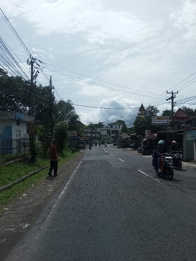 This image depicts a two-lane asphalt road scene in the city of Sandamal Eliya, Sri Lanka, under an overcast sky with scattered clouds. The road, appearing damp in sections, extends into the distance, with a white dividing line visible.
On the left side of the road, a male individual is standing on a dirt and gravel shoulder, facing away from the camera and slightly towards the road. He is wearing a brown patterned long-sleeved shirt and dark trousers. Behind him are dense green trees and bushes, partially obscuring a white building with a blue door and a black metal fence. A prominent utility pole with numerous overhead wires runs along this side of the road.
On the right side of the road, two individuals on motorbikes are visible in the midground, traveling away from the camera. The lead rider wears a light blue helmet and a dark jacket, operating a dark-colored scooter. The second rider, trailing slightly, wears a pink/red helmet and a dark jacket, also on a dark-colored scooter. Further down this side, various buildings with visible red-tiled roofs and a distinctive building with a reddish-brown turret-like structure are present. A shop sign displaying "MY TOKO" in large letters, with smaller Indonesian text below it, is mounted on a pole or building. Multiple utility poles with overhead wires are also present on the right. In the far distance, several other motorbikes and at least one car are visible on the road.