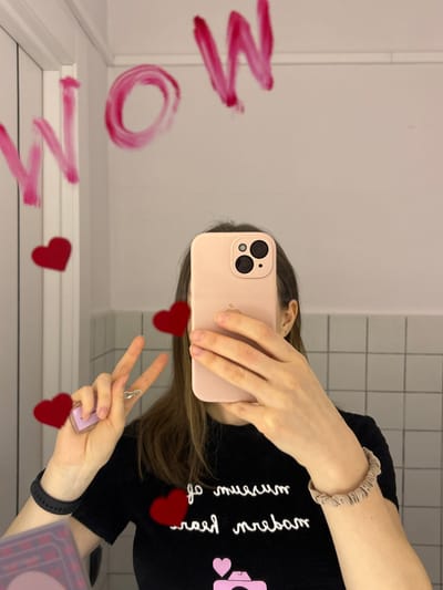 The image depicts a mirror selfie taken indoors in Novosergievka, Russia. The upper torso and head of an individual with long brown hair are reflected in a mirror, their face obscured by a light pink-cased smartphone held in their right hand. The smartphone features a triple-lens camera array and an Apple logo is visible on its back. The individual is wearing a black t-shirt with white text, reflected backward as "muzeum of modern reert," and a pink camera icon with a heart. A black fitness tracker is worn on the left wrist and a light beige scrunchie on the right wrist. The individual's left hand forms a peace sign and holds a small, light purple translucent object with a silver ring. The word "WOW" is written in smudged pink material, resembling lipstick, across the upper portion of the mirror. Several digitally added or sticker-like red heart shapes are visible on the mirror, positioned around the reflected person. The background reflected in the mirror consists of a light-colored wall, with a section of light grey square tiles visible behind the subject.