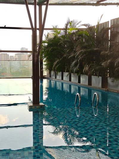 This is a medium shot of a swimming pool, likely on a rooftop in Mumbai, India. The pool is lined with small, dark blue and light blue tiles, creating a checkerboard pattern. Two silver handrails for the pool ladder are visible, emerging from the water. The water itself is a deep turquoise, reflecting the surrounding environment.

Behind the pool, a row of large, rectangular planters filled with lush, dark green palm trees creates a tropical ambiance. Beyond the palm trees, a metal fence is visible. In the background, a cityscape with numerous tall buildings and apartments suggests the urban setting. The sky is bright and overcast, hinting at a possibly humid or cloudy day, typical of Mumbai's climate. Sunlight glints off the pool's surface and reflects in the water, creating interesting patterns.

In the distance, a very small bird is perched on a horizontal beam in the foreground, near the left edge of the frame. The overall scene evokes a sense of relaxation and a private oasis within a bustling city. There is no visible text in the image. The time of day appears to be late morning or early afternoon, given the brightness of the sky and the angle of the light.