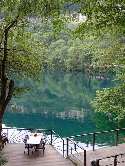 This image captures a serene outdoor dining area by a tranquil lake, situated in the city of Verkhnyaya Balkariya, Russia. A wooden deck, furnished with a table and four chairs, overlooks a body of turquoise water. The lake's surface reflects the lush greenery of the surrounding trees and the rocky cliffs in the distance. The water is calm, with subtle ripples indicating a gentle breeze. Overhanging branches of trees frame the scene, adding to the natural beauty. A date and time stamp, "26.07.2026 15:39," is visible in the lower right corner. The environment is predominantly natural, with a focus on the pristine landscape and the simple, functional dining setup.