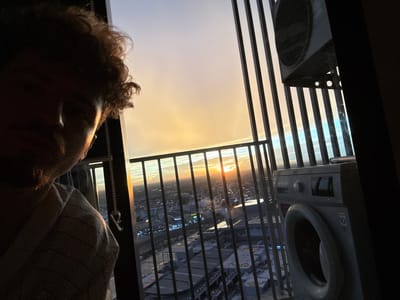 The image shows a person in the foreground, with their head and shoulders visible, facing away from the camera and towards a large window. The person has curly hair and is wearing a striped shirt. Through the window, a city skyline is visible at dusk, with the sun setting in the distance, casting a warm glow over the scene. The city appears to be very dense with tall buildings.
In the foreground, to the left of the person, are two appliances. One is a washing machine, and the other appears to be an air conditioning unit. These are positioned behind a set of vertical bars, suggesting they might be on a balcony or an external utility area. The overall setting appears to be an urban environment, likely a high-rise building, given the perspective of the city below and the presence of these appliances. The mood of the image is somewhat contemplative, with the person gazing out at the city and the sunset. The lighting suggests it's the golden hour, just before nightfall. There is no visible text in the image.