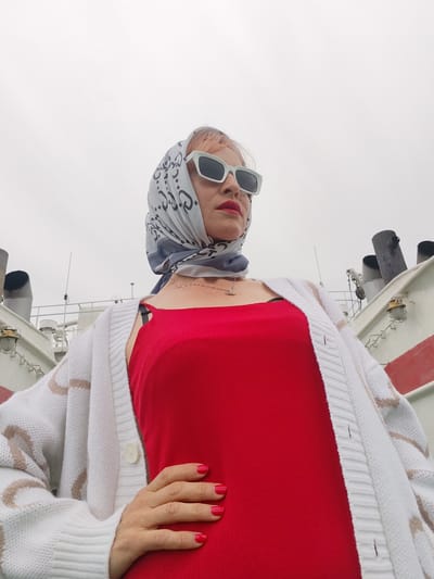 A woman with pink-tinted hair, wearing a red slip dress, a white cardigan with beige stripes, a headscarf with a gray pattern, and white rectangular sunglasses, poses on a boat. She has bright red nail polish on her fingernails. Her left hand is on her hip. The background shows parts of a boat, including smokestacks and railings, under a cloudy sky. The scene suggests an overcast day. The image appears to be a self-portrait taken from a low angle. No text is visible in the image. The location context is Bereg, Russia.