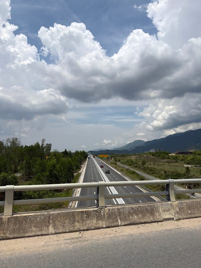 An eye-level, medium outdoor shot captures a highway vista in Vietnam under a cloudy sky. The highway stretches into the distance, with lanes demarcated by white lines. A few cars are visible on the road. To the left, a dense line of green trees borders the highway, creating a stark contrast with the gray concrete barrier and railing in the foreground, which partially obscures the view. In the background, a range of mountains with lush green slopes rises against the sky, dotted with a few white clouds. The scene is devoid of any people. The image is likely taken from a bridge or overpass, as indicated by the railing and the perspective.