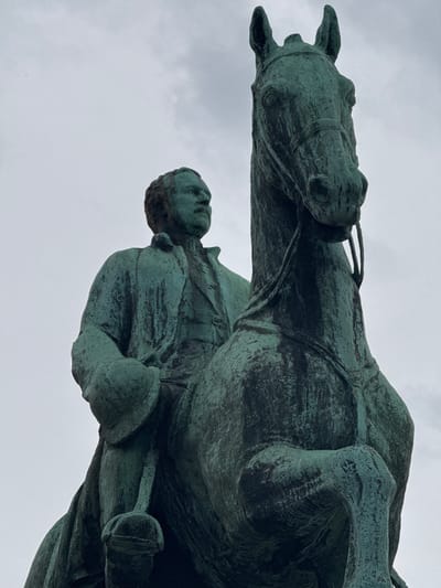 An equestrian statue of King Leopold I, made of bronze, stands against a cloudy sky in Brussels, Belgium. The statue depicts the king, wearing formal attire, seated on a horse. The bronze has developed a verdigris patina, giving it a greenish hue. The viewpoint is from below, looking up at the statue, emphasizing its scale and presence. The horse's head is turned slightly to the left, while the king's gaze is directed forward. The background is an overcast sky with visible cloud formations.