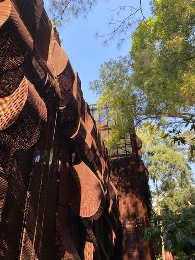 The image captures a unique architectural structure in the city of Ciwaruga, Indonesia. The structure is made of rusted metal panels that form an undulating, textured facade. These panels have a perforated pattern, creating a play of light and shadow. The perspective is from below, looking up towards the sky and through the structure. Green trees and foliage are visible around and above the building, suggesting a natural or park-like setting. The sky is clear and blue, indicating a sunny day. There are no people visible in the image. The overall impression is one of an artistic and organic-looking building integrated with its natural surroundings.