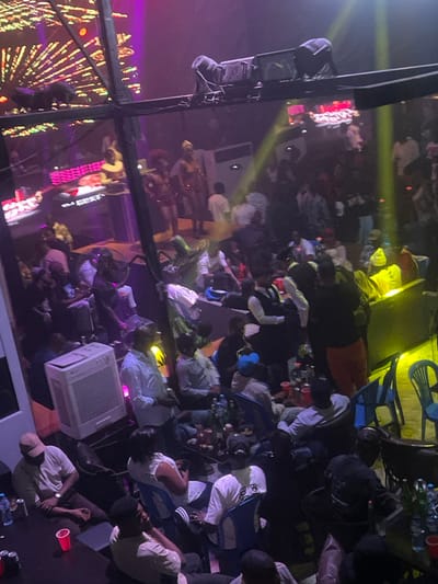 A crowded indoor scene shows many people sitting on various surfaces, perhaps the floor. Some individuals are wearing white shirts. Drinks in red cups are scattered throughout. An air conditioning unit sits among the crowd. Stage lighting illuminates sections, with reflections on a shiny surface adding complexity. Part of a stage and a lighting rig are visible. The overall impression is of a casual gathering in a club or event space.
