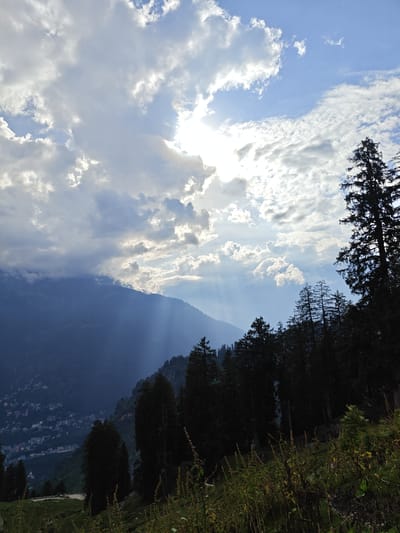 The image captures a natural landscape with a village nestled in the valley, likely in Chhyal, India. The foreground features tall, dark evergreen trees and dry grass. A winding road is visible in the lower left corner. In the mid-ground, the slopes of a mountain are covered with a dense forest, with a cluster of buildings forming a village spread across the lower portion of the mountain. The background is dominated by a hazy, dark mountain range. The sky above is a mix of blue sky and heavy clouds, with rays of sunlight breaking through, creating a dramatic effect. The overall impression is one of a remote, mountainous region experiencing a dynamic interplay of light and weather.