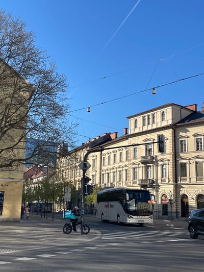 The image shows a street scene in Ljubljana, Slovenia. A white tour bus is parked on the right side of the street, partially blocking a zebra crossing. A delivery person on a scooter with a "Wolt" bag is in the foreground. The background features a multi-story building with ornate windows and architectural details. Power lines are visible against a clear blue sky. A contrail from a plane can be seen in the upper left corner of the frame. A tree with bare branches occupies the lower left portion of the image, partially obscuring other buildings and people on the sidewalk.