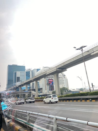 The image depicts a street scene in South Jakarta, Indonesia, likely during the day. The perspective is from inside a vehicle, looking out. A raised, concrete structure, possibly an elevated highway or train line, dominates the upper left portion of the frame. Underneath this structure, multiple lanes of traffic are visible.
Several cars are driving on the road, including a white SUV in the foreground and a black and white sedan further down. The road is wet, suggesting it has recently rained or is currently raining, and the sky is overcast and bright white, further indicating cloudy or rainy weather.
Tall modern buildings line the right side of the road. One building has "KEMENTERIAN KEUANGAN INDONESIA" written on its side, identifying it as the Ministry of Finance of Indonesia. Another building is labeled "TOWER KEMANG". Advertisements are displayed on screens on some of the buildings.
The scene conveys a sense of urban, busy city life, with traffic and large buildings characteristic of a major metropolitan area. The lighting is diffused, suggesting overcast skies. There are no visible people in the image, but the presence of cars implies human activity. The overall mood is neutral, focusing on the urban landscape and traffic.